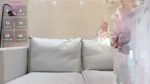 Media: Video of a white sectional sofa in a modern, bright room, with a blurred person in the background wearing a pink dress, and a digital display showing a calendar.