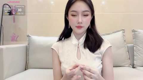 Media: Video of an Asian woman with straight black hair, fair skin, wearing a white cheongsam with a high collar, sitting on a light-colored sofa. Background features a beige wall with a digital clock and heart-shaped decorations.