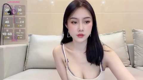 Media: A video of an Asian woman with straight black hair, fair skin, and red lipstick, wearing a white spaghetti strap top, sitting on a cream couch with beige pillows.