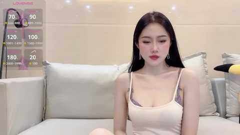 Media: Video of an East Asian woman with long black hair, wearing a beige tank top and purple lace bra, seated on a beige couch in a modern, minimalist room.