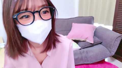 Media: Video of a young Asian woman with straight, shoulder-length brown hair, wearing black-framed glasses, a white face mask, and a light pink blouse, sitting on a gray couch with pink pillows in a modern living room.