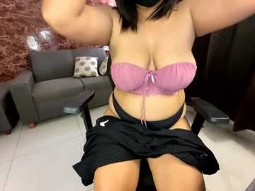 Media: Video of a curvy woman with light skin, wearing a pink lace bra and black skirt, sitting in a chair in a modern living room with a gray sofa and marble wall.