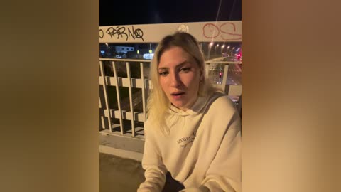 Media: Video of a blonde woman with a surprised expression, wearing a cream hoodie, standing on a bridge at night with graffiti and city lights in the background.