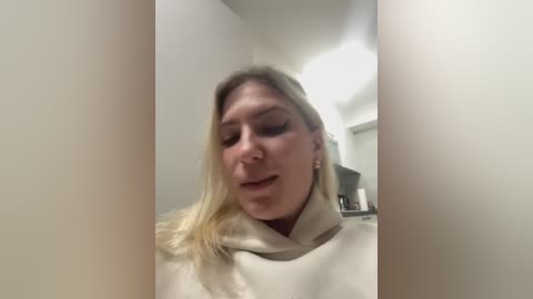 Media: Video of a smiling woman with long blonde hair, wearing a beige hoodie, captured from a selfie angle with blurred background showing a kitchen.