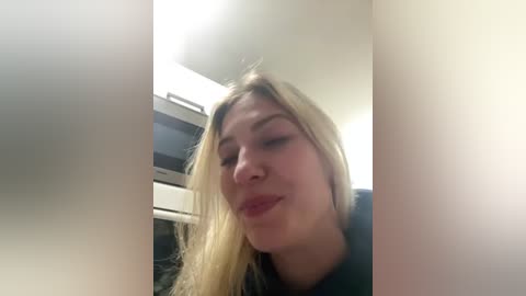 Media: Video of a blonde woman with fair skin, wearing a black top, making a playful face in a modern kitchen with white cabinets and a microwave.