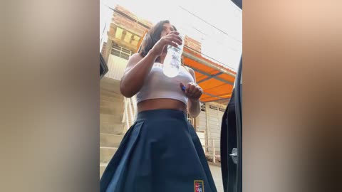 Media: Video of a young woman in a white crop top, blue pleated skirt, and a school bag, drinking water outside, with a brick building and orange awning in the background.