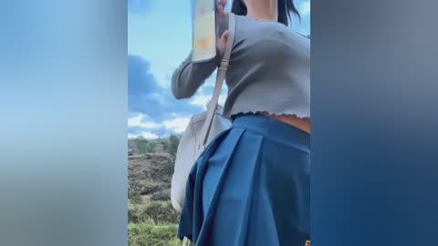 Media: Video of a young woman with a medium build, wearing a cropped gray sweater and a blue pleated skirt, taking a selfie outdoors with a cloudy sky and greenery in the background.