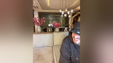 Media: Video of a smiling man in a black hat and jacket, sitting on a beige couch in a modern, dimly lit living room with a large window, potted red flowers, and a chandelier.