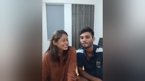 Media: Video of a young South Asian couple, smiling, sitting on a bed in a room with white walls and a window with brown curtains. The woman wears a brown sweater, and the man a navy polo shirt.