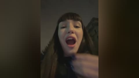 Media: A video of a young woman with long, straight dark hair and blunt bangs, open-mouthed and looking upwards, possibly in a playful or surprised expression. The background is blurred and dimly lit.