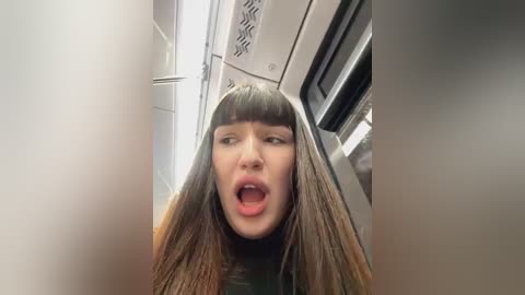 Media: A video of a young woman with long, straight brown hair, wearing a black top, standing in a modern, metallic elevator with a surprised expression and open mouth.