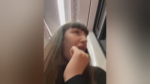 Media: A blurry video of a young woman with long, straight hair, wearing a black top, looking thoughtful and holding her chin, in an elevator with beige walls and a patterned carpet.