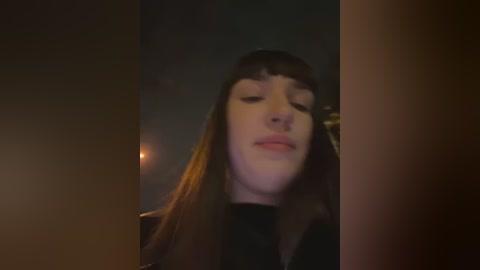 Media: Video of a young woman with long, straight brown hair and bangs, wearing a black top. She has a slight smile, eyes closed, and is indoors in a dimly lit room with a blurred background.