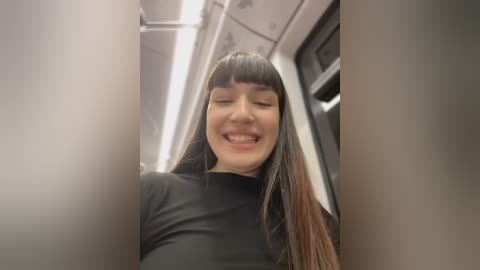 Media: Video of a young woman with straight, dark brown hair and bangs, wearing a black top, smiling and laughing in a subway car.