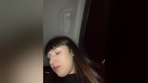 Media: Video of a young woman with long brown hair and bangs, wearing black clothing, sitting in a dimly lit car with a dark interior, eyes closed and head tilted slightly, appearing relaxed.