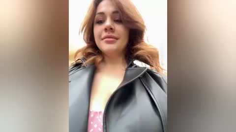 Media: Video of a fair-skinned woman with wavy, shoulder-length auburn hair, wearing a black leather jacket and a pink shirt with white polka dots, smiling confidently.