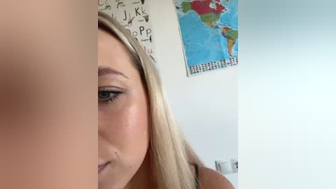 Media: Video of a close-up of a young woman with light skin, long blonde hair, and a slight pout. Background features a map of the world and a white wall.