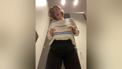 Media: Video of a woman in a white, striped sweater, black leggings, and brown hair, captured from a low angle, laughing with her mouth open, in a modern, beige-walled hallway with recessed lighting.