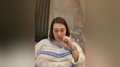 Media: Video of a young woman with shoulder-length brown hair, wearing a white and blue striped sweater, sitting indoors with her chin resting on her hand, looking introspective.