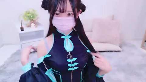 Media: Video of a young Asian woman with long black hair styled in double buns, wearing a blue and teal cosplay dress, a pink face mask, and a teal bow. She stands in a minimalist, light-colored room with a white wall and a fluffy grey rug.