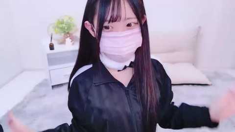 Media: Video of an Asian woman with long black hair, wearing a white face mask, black shirt, and a black choker, seated on a gray carpet in a minimalist, white-walled bedroom with a white bed and a white nightstand.