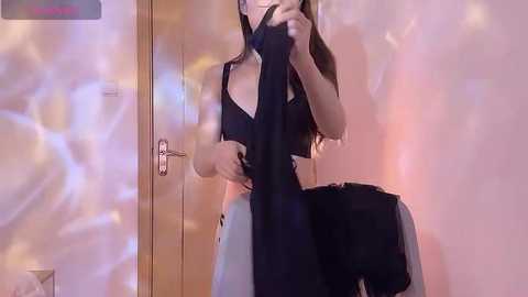 Media: A video captures a light-skinned woman with long brown hair, wearing a black crop top and white pants, taking a selfie in a dimly lit room with a blurred, colorful background.