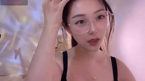 Media: Video of an East Asian woman with fair skin, wearing clear glasses and a black tank top, adjusting her hair in a soft, diffused room with a blurred abstract painting background.