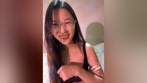 Media: Video of a young Asian woman with long black hair and glasses, smiling, wearing a black tank top, in a dimly lit room.