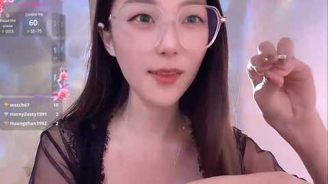 Media: Video of an Asian woman with long black hair, fair skin, and clear glasses, wearing a sheer black lace top. Background shows blurred indoor scene with social media notifications.