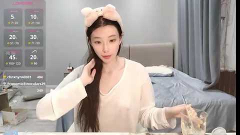 Media: A video of an Asian woman with long black hair, wearing a white robe and bunny ears, sitting at a table in a bedroom.