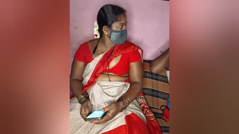 Media: Video of an Indian woman in a red and beige sari, wearing a face mask, seated on a bed with a pink pillow, holding a smartphone, in a dimly lit room.