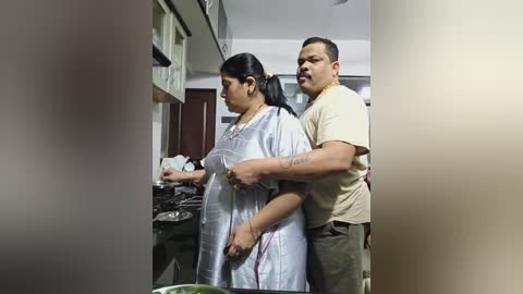 Media: Video of a man with glasses restraining a woman in a kitchen. Woman wears a white apron, man in beige t-shirt. Background shows cabinets and cluttered countertops.