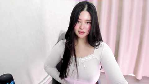 Media: Video of an East Asian woman with long black hair, fair skin, wearing a white lace-trimmed long-sleeve top, seated indoors with pink curtains and a white wall background.