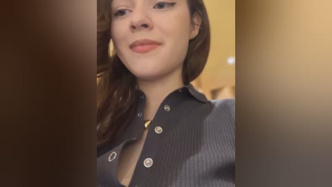 Media: A video of a fair-skinned woman with wavy brown hair, wearing a dark blue, ribbed, button-up shirt, standing in a dimly lit room with a blurred background.