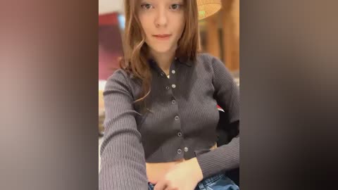 Media: Video of a fair-skinned, light-haired woman with a slim build, wearing a grey ribbed cardigan and blue jeans, sitting indoors with blurred background.