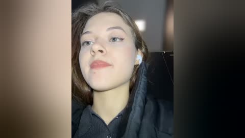 Media: Video of a young Caucasian woman with fair skin, brown hair, and light makeup, wearing a black jacket, seated indoors, with a blurred background.