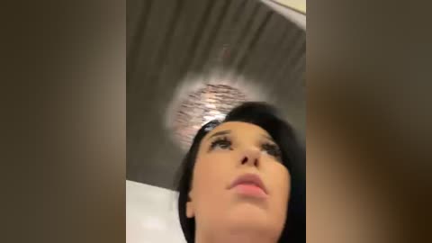 Media: A video of a woman with light skin, dark hair, and a shiny, metallic halo hovering above her head. She has a serious expression and is indoors with a gray ceiling.