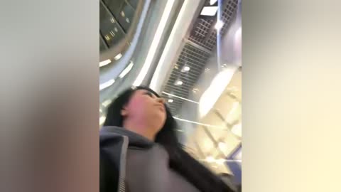 Media: Video of a young woman with dark hair and glasses, wearing a gray hoodie, looking upward in a modern, bright elevator with glass walls and LED lights.