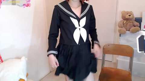 Media: Video of a young woman in a black schoolgirl outfit with white bow, standing in a room with a teddy bear, wooden chair, and white wall.