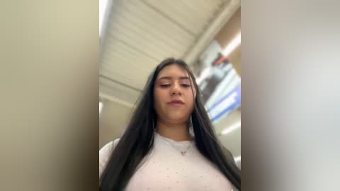 Media: Video of a young Latina woman with long, straight black hair, wearing a white T-shirt and a silver necklace, standing in an indoor, bright, modern setting with a metallic ceiling and blurred background.