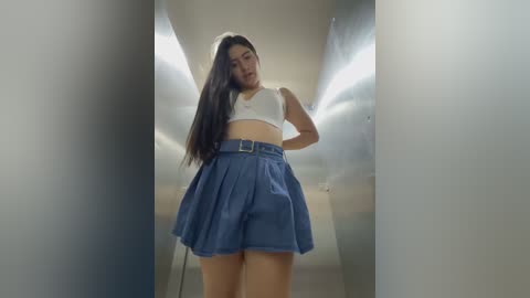 Media: A video of a young Asian woman with long black hair, wearing a white crop top and high-waisted blue pleated skirt, standing in a narrow, dimly lit hallway.