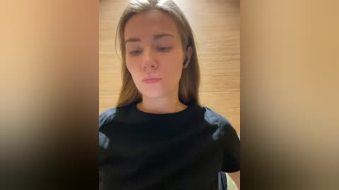 Media: Video of a young Caucasian woman with fair skin and long, straight brown hair, wearing a black t-shirt, standing against a wooden wall. She appears contemplative.