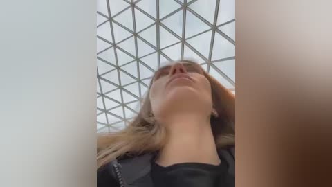 Media: Video of a blonde woman with light skin and straight hair, looking up at a grid-like skylight ceiling. She wears a black jacket. The image is low-angle, emphasizing her profile.