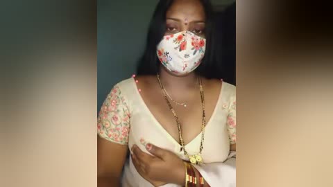 Media: Video of a woman with medium brown skin, long black hair, wearing a floral-patterned mask, traditional white blouse with gold jewelry, and a gold chain necklace, against a blurred indoor background.