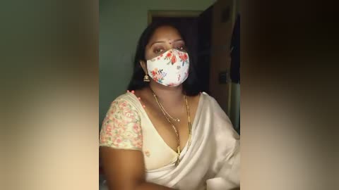 Media: Video of a woman with medium brown skin, wearing a floral-patterned face mask, white sari, and gold necklace, standing in a dimly lit, indoor setting with a doorway visible behind her.