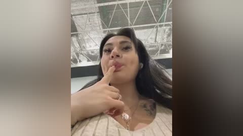 Media: Video of a Latina woman with long, straight black hair, medium skin tone, and a tattoo on her left shoulder. She's indoors, holding a finger to her lips, wearing a beige sweater.