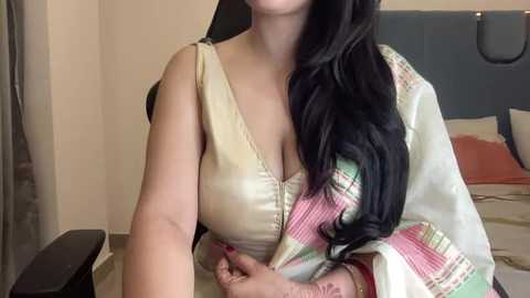 Media: Video of a South Asian woman with long black hair, wearing a beige sleeveless top with a low neckline, revealing cleavage. She sits on a bed with a blue headboard, surrounded by beige walls and a dark wooden nightstand.