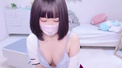 Media: Video of an Asian woman with bobbed hair and a mask, wearing a white lace bra and white gloves, kneeling in a pastel bedroom with a bed and dresser.