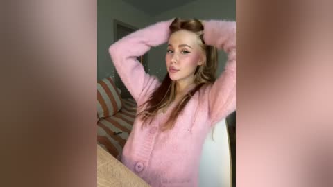 Media: A video of a young woman with fair skin, long auburn hair, and a slender physique, wearing a pink fuzzy cardigan, sitting on a striped couch, arms raised. Background shows a beige wall and a door.