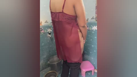 Media: Video of a woman with medium skin tone, wearing a sheer red lingerie dress, standing in a bathroom with a pink stool and floral wallpaper.